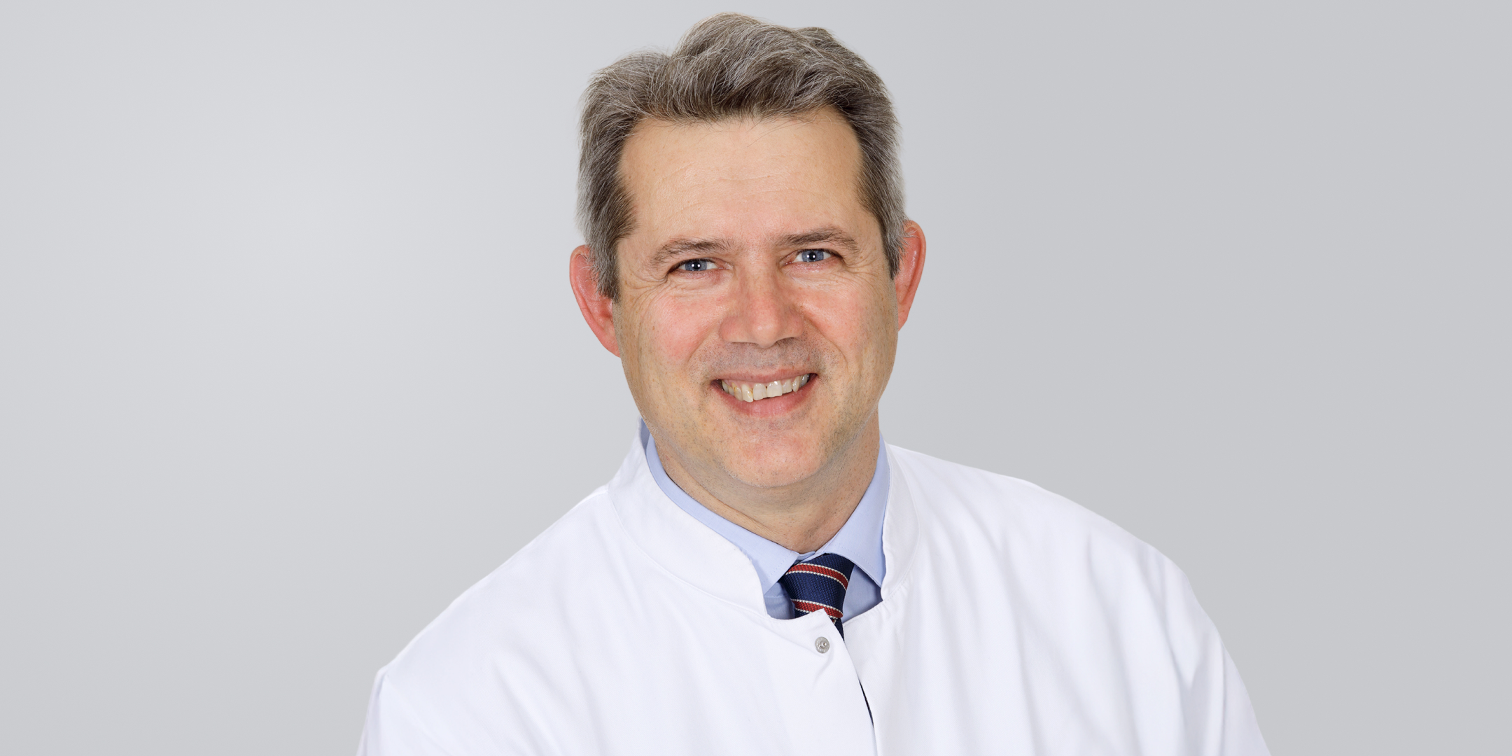 Lächelnder Mann mit kurzen Haaren und weißem Kittel vor grauem Hintergrund.