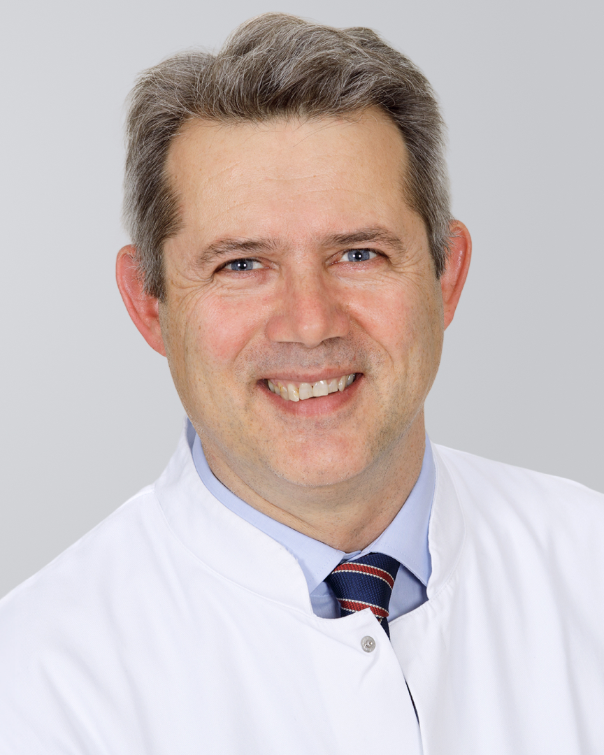 Lächelnder Mann mit kurzen Haaren und weißem Kittel vor grauem Hintergrund.
