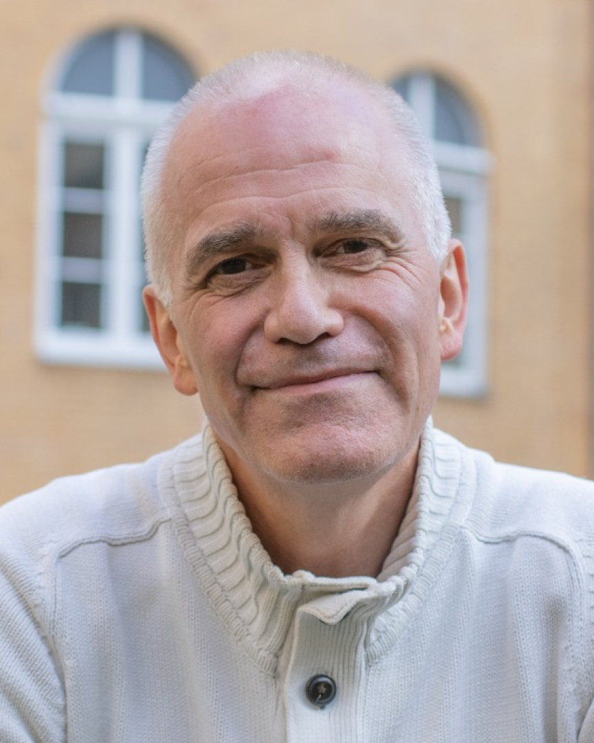 Ein älterer Herr vor einer Hausfassade mit vielen Fenstern, der in die Kamera lächelt