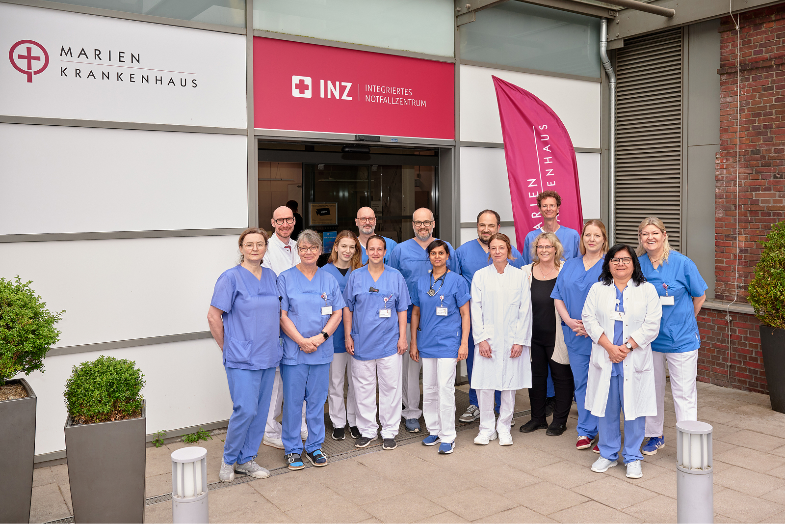 Gruppe von medizinischem Personal in blauer Arbeitskleidung und weißen Kitteln vor einem Eingang mit einem roten Schild und Fahnen.
