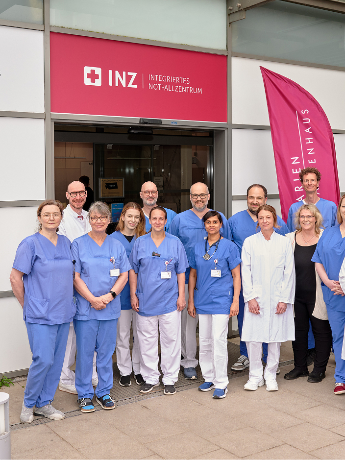 Gruppe von medizinischem Personal in blauer Arbeitskleidung und weißen Kitteln vor einem Eingang mit einem roten Schild und Fahnen.