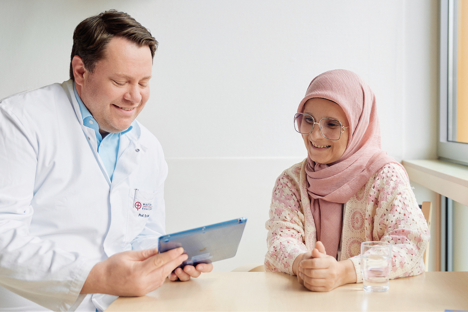 Arzt im weißen Kittel hält lächelnd ein Tablet in der Hand, er zeigt es einer Patientin, die neben ihm sitzt.
