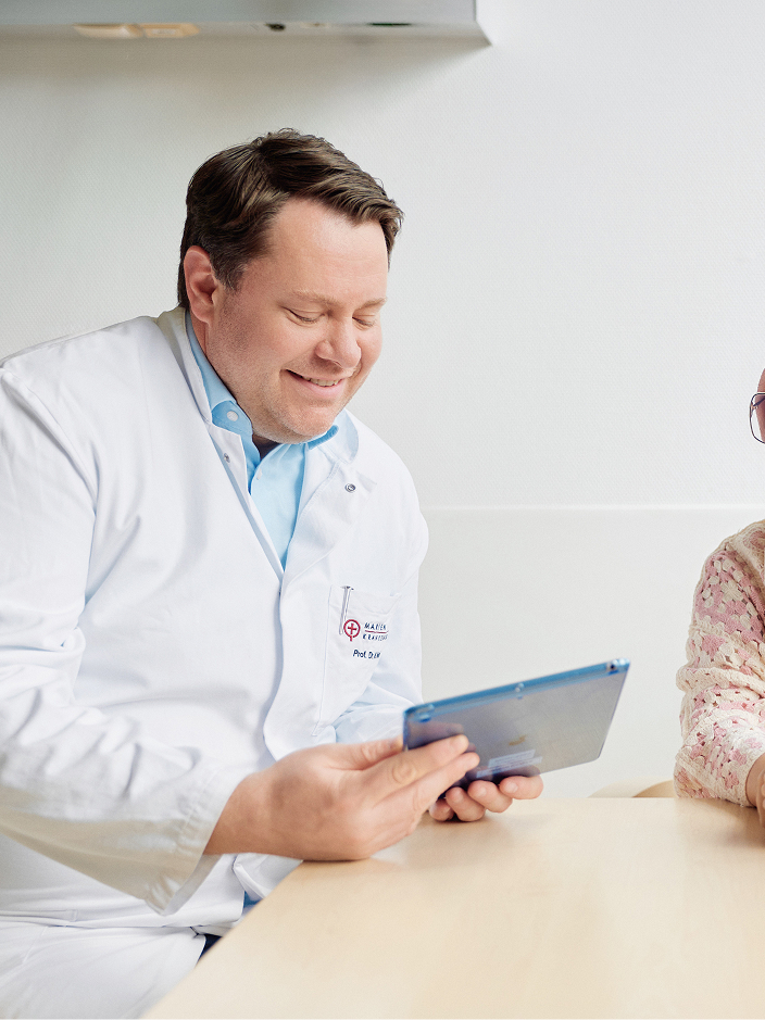 Arzt im weißen Kittel hält lächelnd ein Tablet in der Hand, er zeigt es einer Patientin, die neben ihm sitzt.