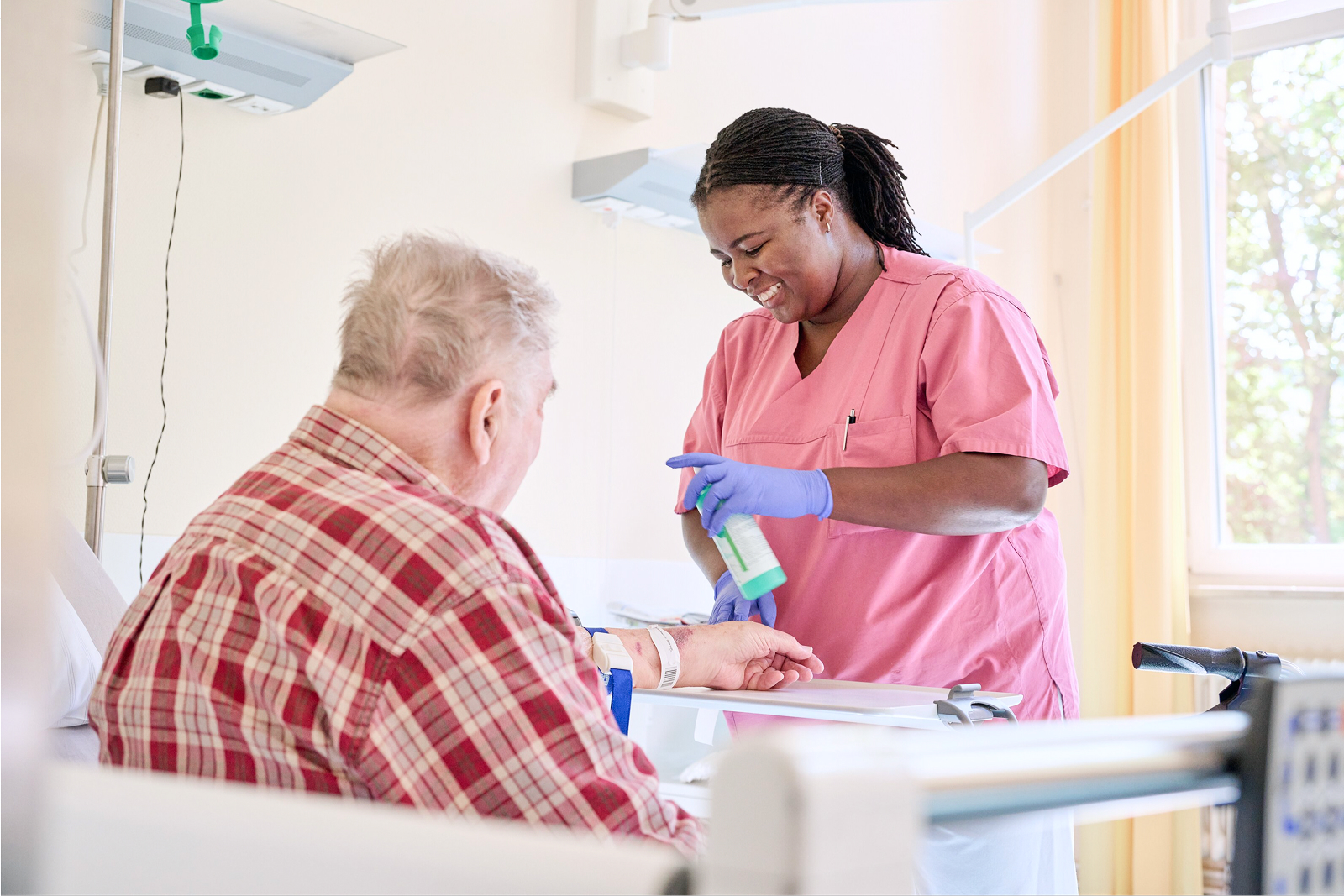 Eine Pflegekraft in rosafarbener Arbeitskleidung behandelt einen älteren Patienten am Handgelenk.