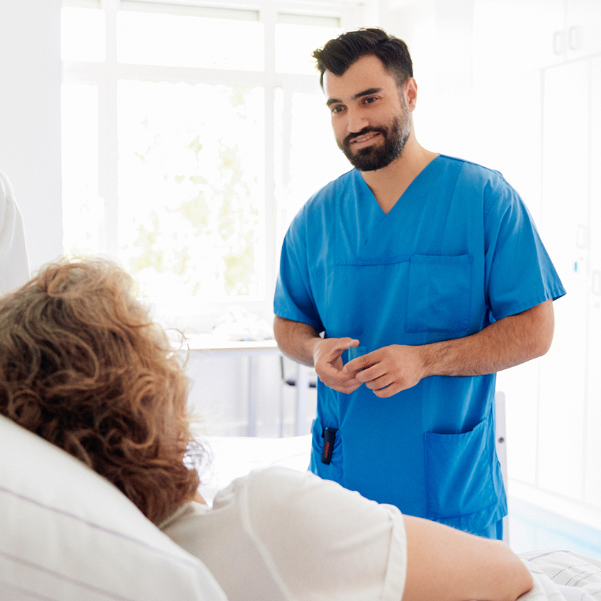 Ein Arzt in blauer Arbeitskleidung spricht mit einer Patientin, die im Krankenhausbett liegt.