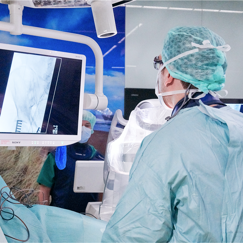 Ein Chirurg in Schutzkleidung betrachtet einen Monitor während eines medizinischen Eingriffs in einem Operationssaal.