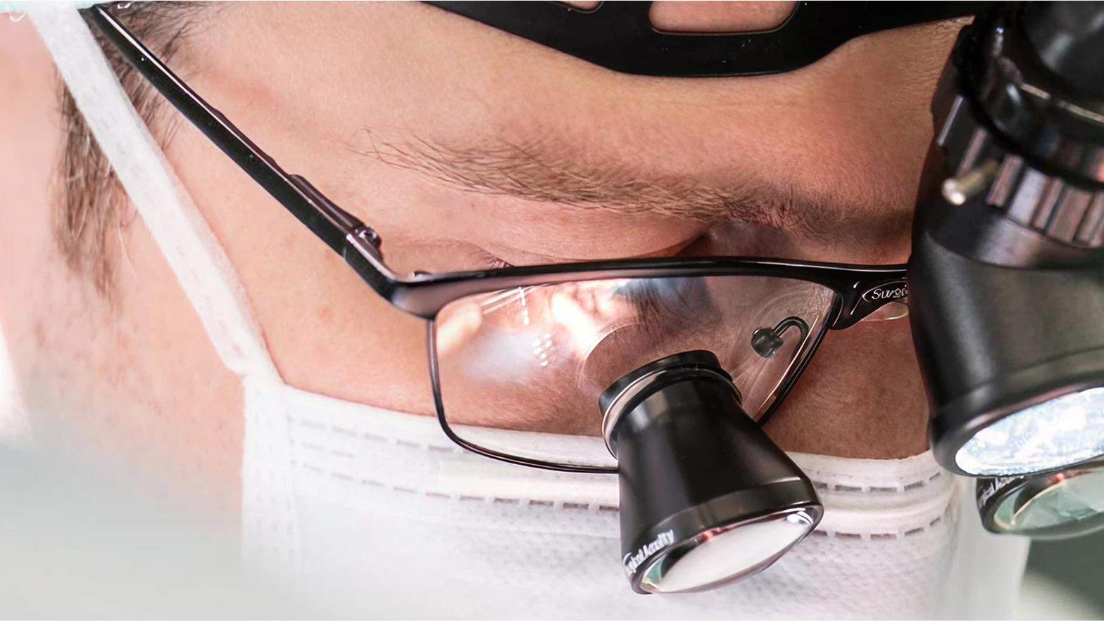 Nahaufnahme eines Gesichts mit OP-Brille und einer Lupe, die Person trägt einen weißen Mundschutz.