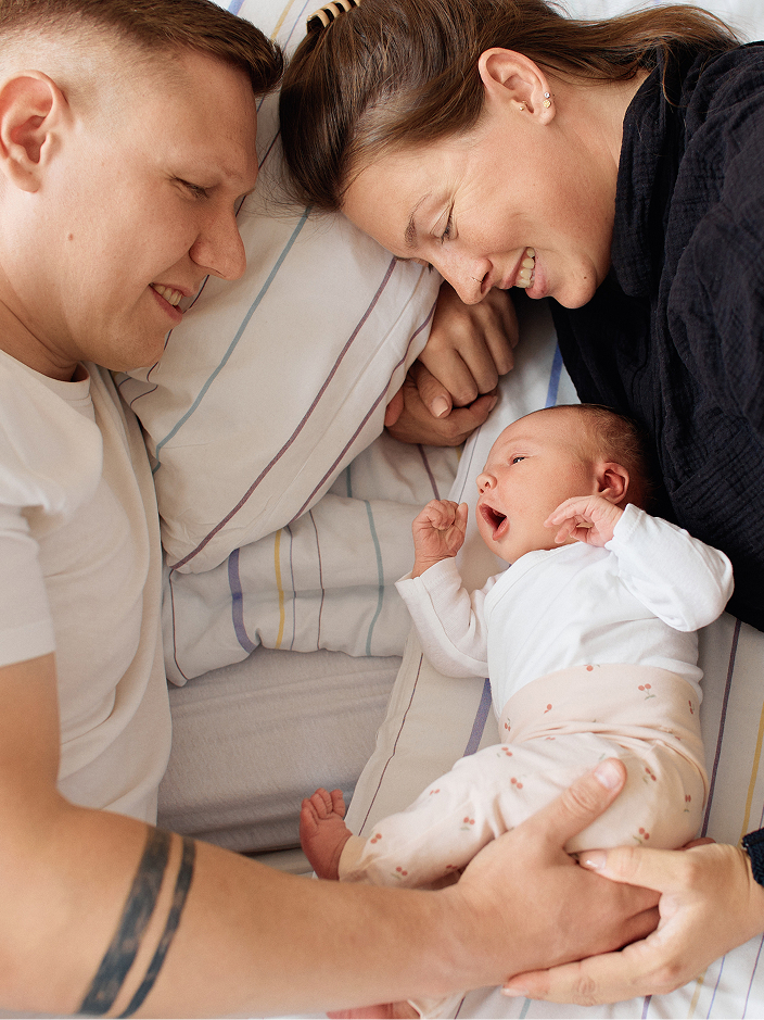 Eine Familie liegt auf einem Bett. Die Eltern schauen liebevoll auf ihr gähnendes Baby, das zwischen ihnen liegt.