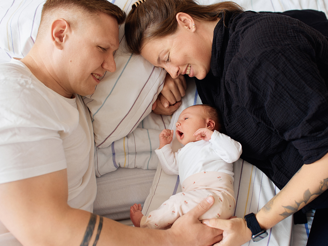 Eine Familie liegt auf einem Bett. Die Eltern schauen liebevoll auf ihr gähnendes Baby, das zwischen ihnen liegt.