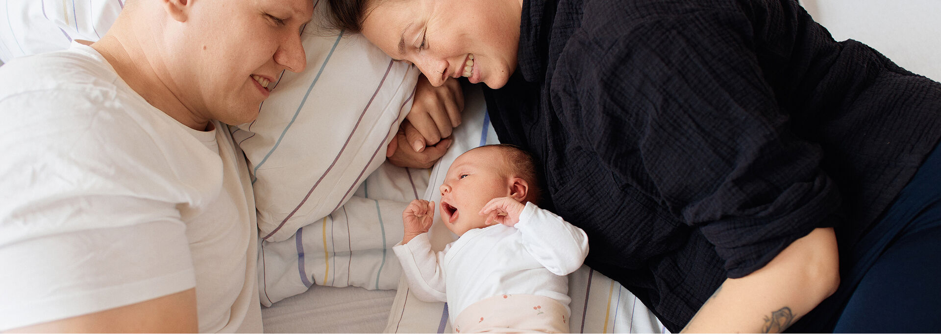 Eine Familie liegt auf einem Bett. Die Eltern schauen liebevoll auf ihr gähnendes Baby, das zwischen ihnen liegt.