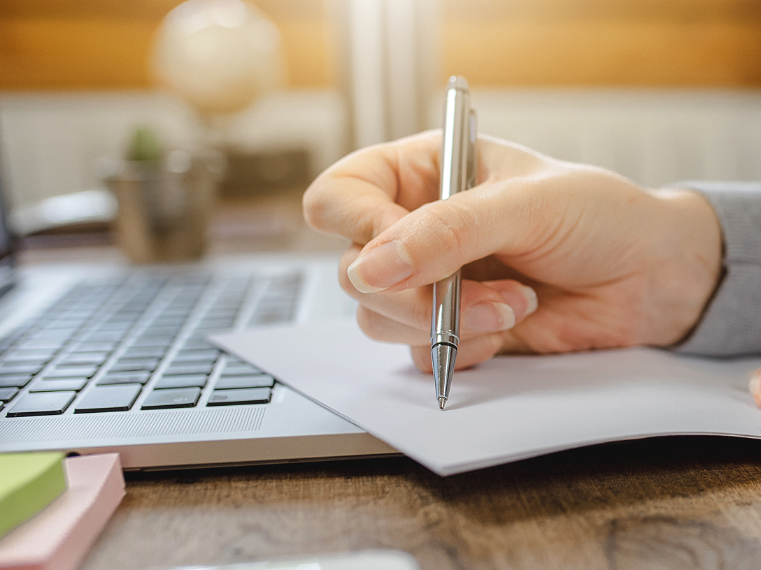 Eine weibliche Hand hält einen Stift und schreibt auf ein Blatt Papier, das auf der Tastatur eines Laptops liegt.
