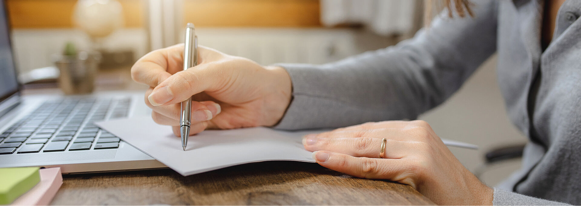 Eine weibliche Hand hält einen Stift und schreibt auf ein Blatt Papier, das auf der Tastatur eines Laptops liegt.