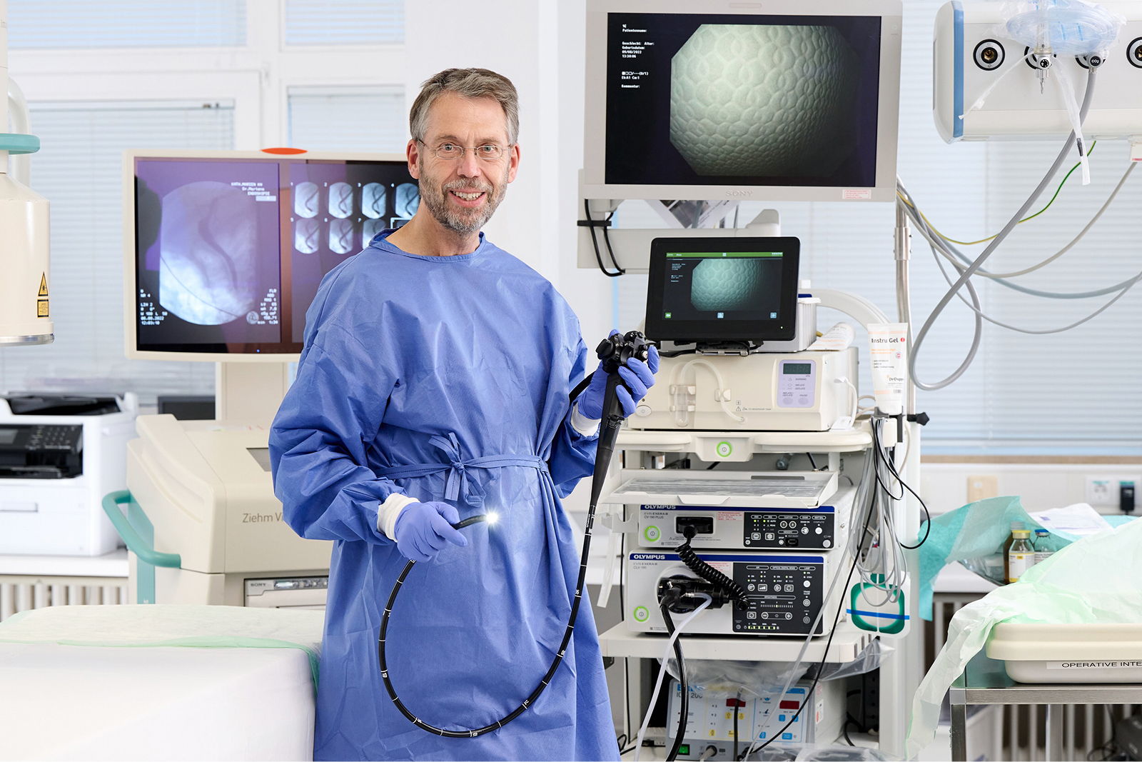 Ein Arzt in blauer Schutzkleidung hält ein Endoskop und steht vor medizinischen Geräten und einem Monitor.