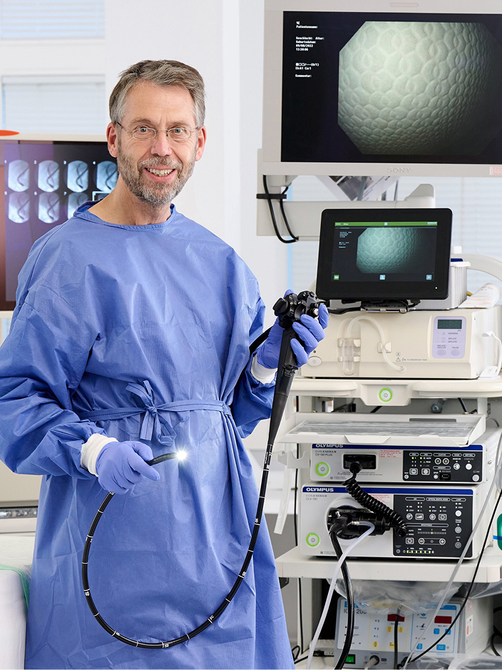 Ein Arzt in blauer Schutzkleidung hält ein Endoskop und steht vor medizinischen Geräten und einem Monitor.