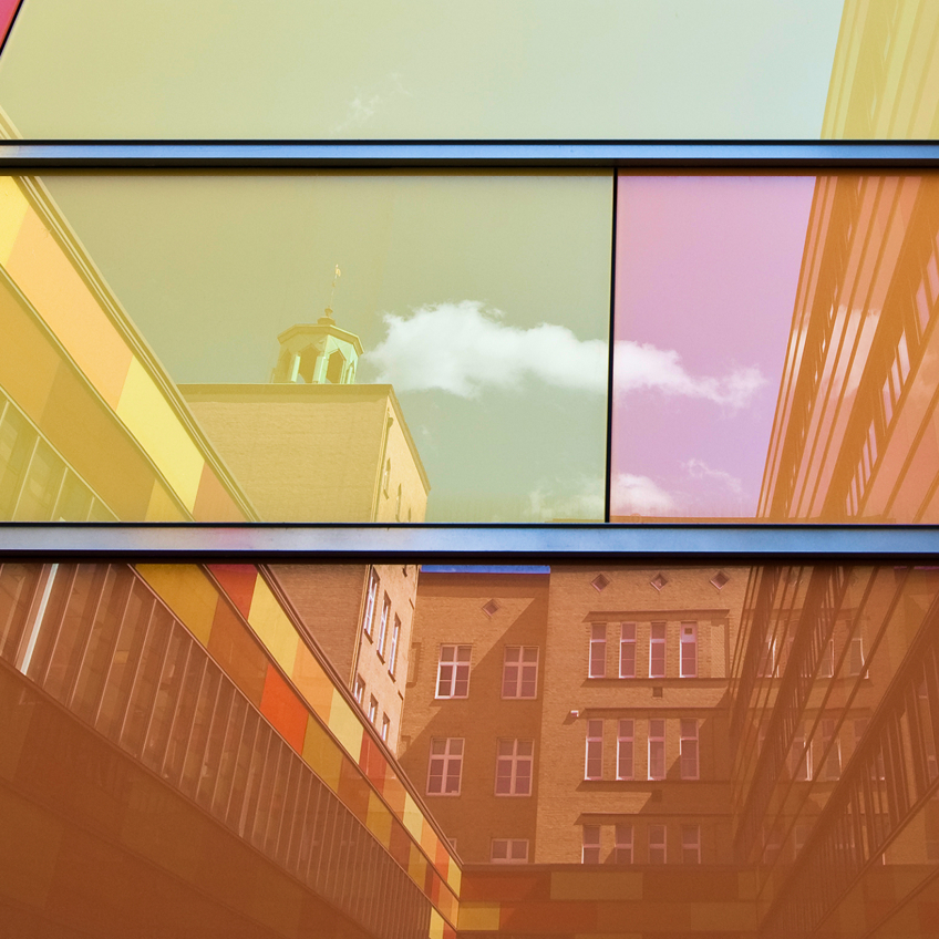 Bunte Glasfassade reflektiert Wolken und angrenzende Gebäude in warmen Farbtönen.