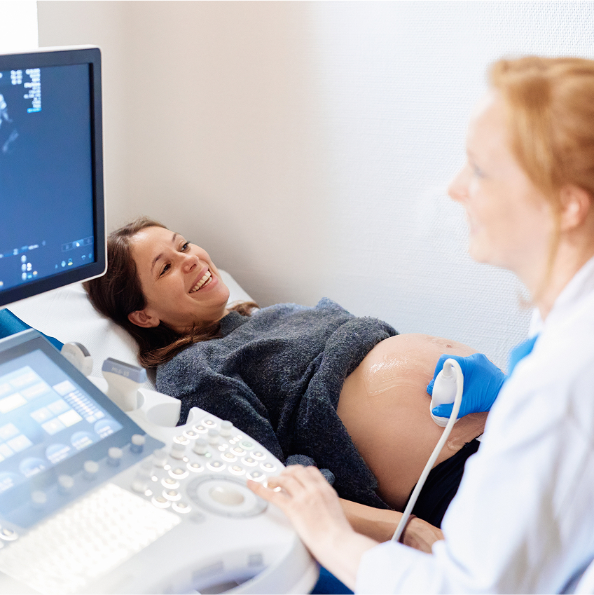 Schwangere Frau lächelt während eines Ultraschalls, eine Ärztin bedient das Gerät und schaut dabei auf einen Monitor.
