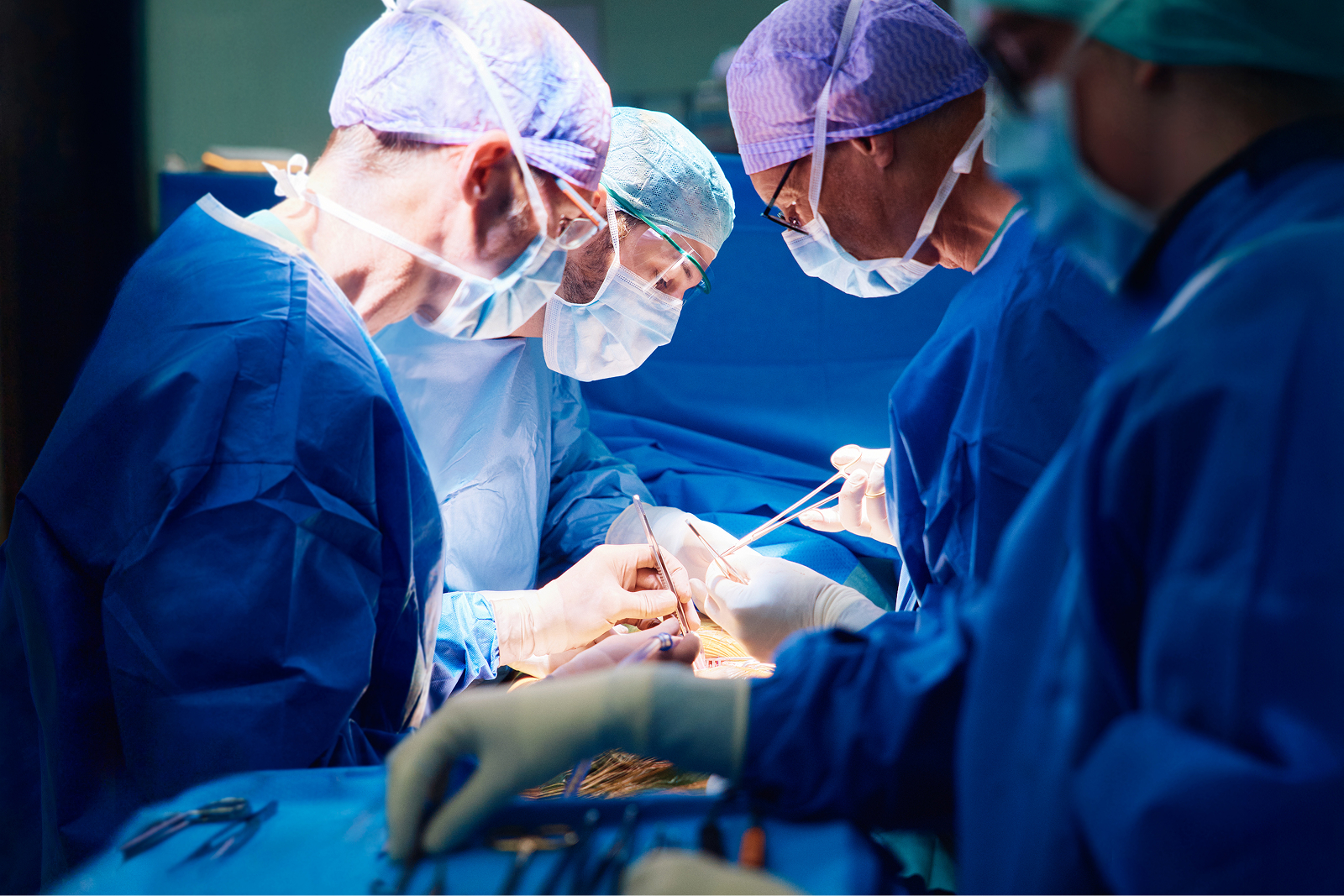 Chirurgen in OP-Kleidung führen eine Operation durch, sie schauen konzentriert auf einen Patienten, der vor ihnen auf dem OP-Tisch liegt.