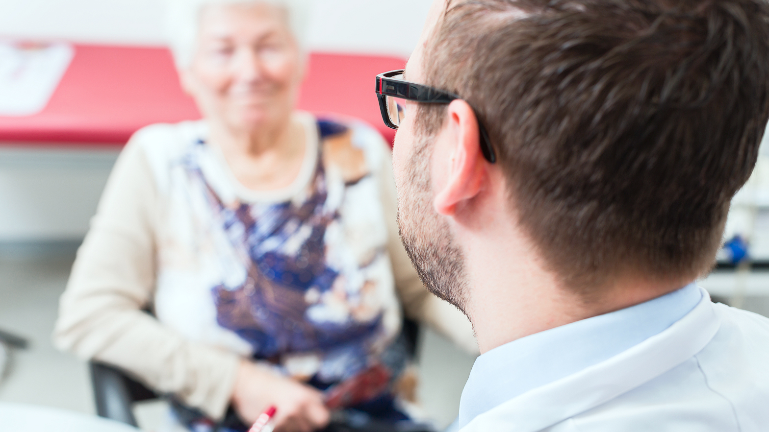 Ein Arzt spricht mit einer älteren Patientin in einer Praxis. Die Patientin lächelt ihn an.