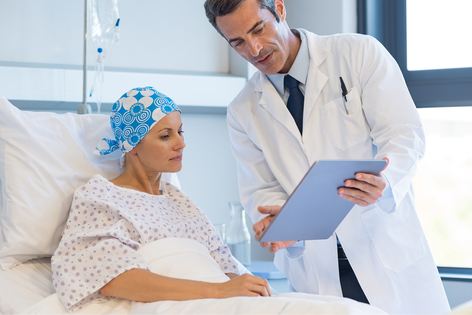 Ein Arzt zeigt einer Patientin im Krankenhaus auf einem Tablet Informationen. Die Patientin sitzt im Krankenhausbett und trägt eine Kopfbedeckung.
