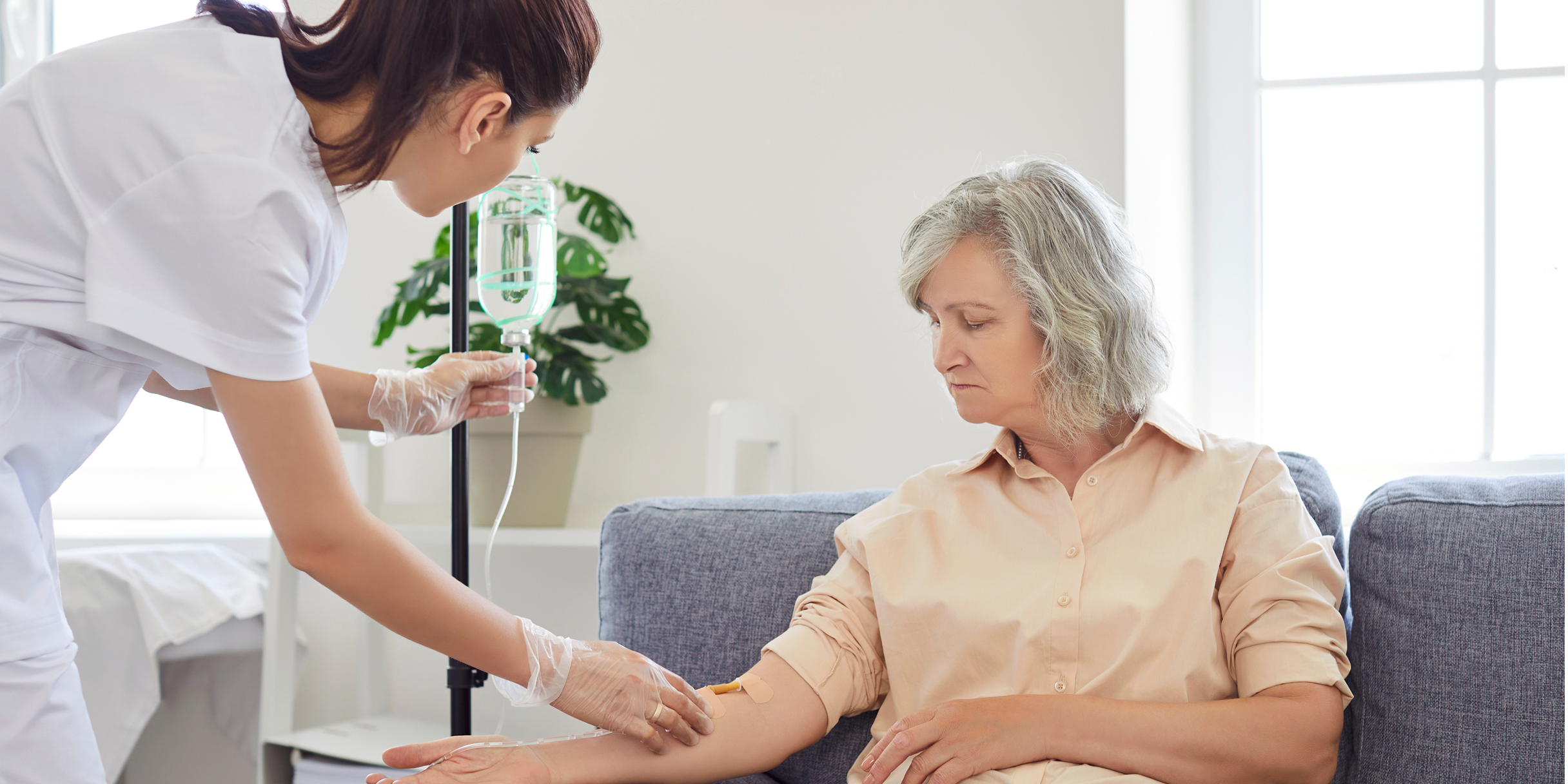 Eine Krankenschwester verabreicht einer älteren Patientin, die in einem hellen Raum auf einem grauen Sofa sitzt, eine Infusion.
