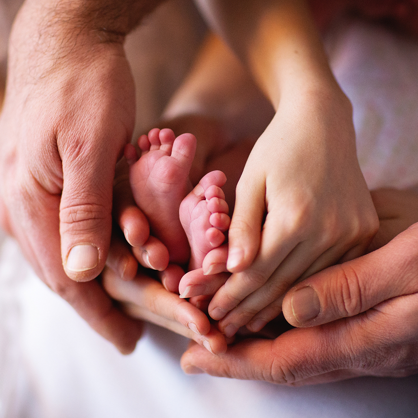 Hände von zwei Erwachsenen umschließen die Füße eines Babys, das auf einer weichen, weißen Oberfläche liegt.