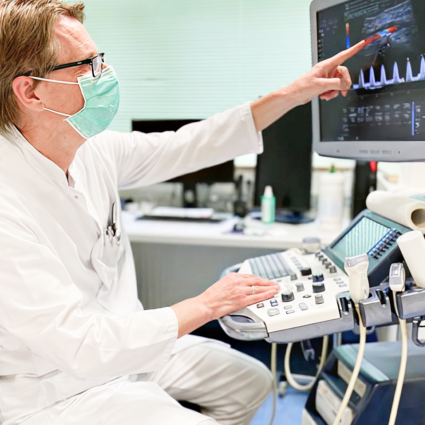 Arzt mit weißem Kittel und Schutzmaske bedient ein Ultraschallgerät und zeigt auf den Bildschirm mit medizinischen Daten.