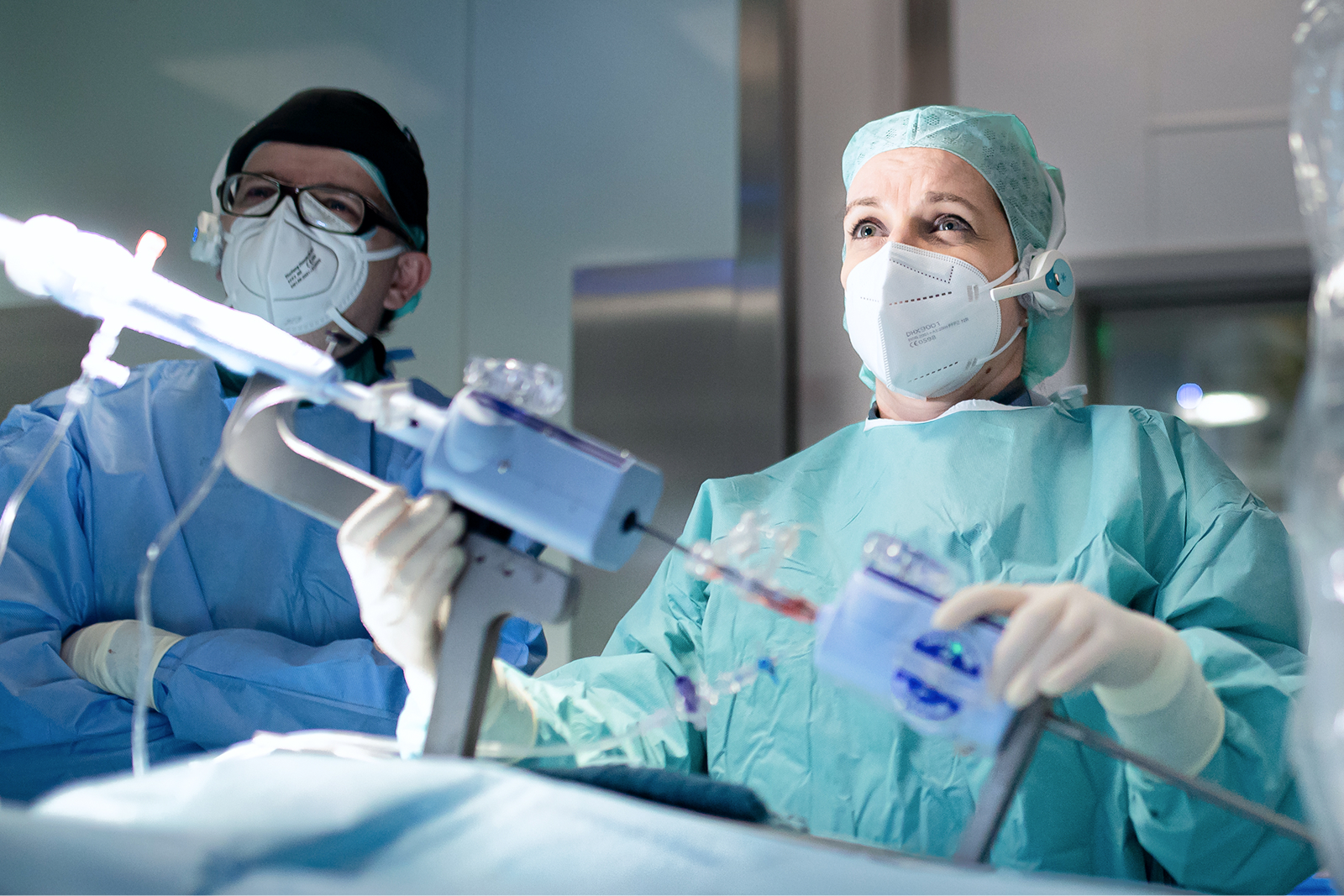 Zwei Chirurgen mit OP-Kleidung und Masken, einer hält ein chirurgisches Instrument, während der andere zuschaut.