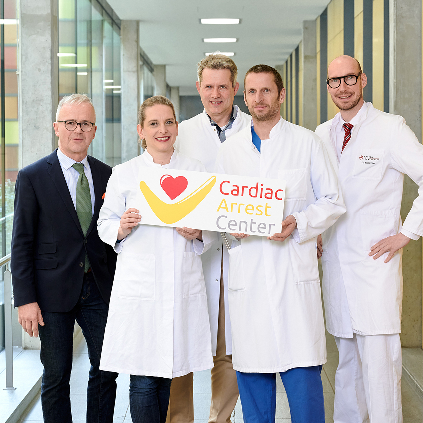 Fünf Personen in weißen Kitteln stehen im Klinikflur, zwei davon halten ein Schild mit der Aufschrift