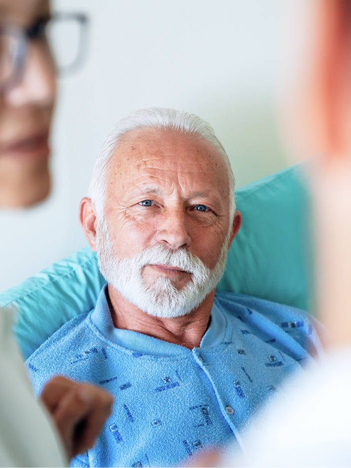 Älterer Mann mit grauem Bart in einem Krankenhausbett, umgeben von zwei medizinischen Fachkräften.