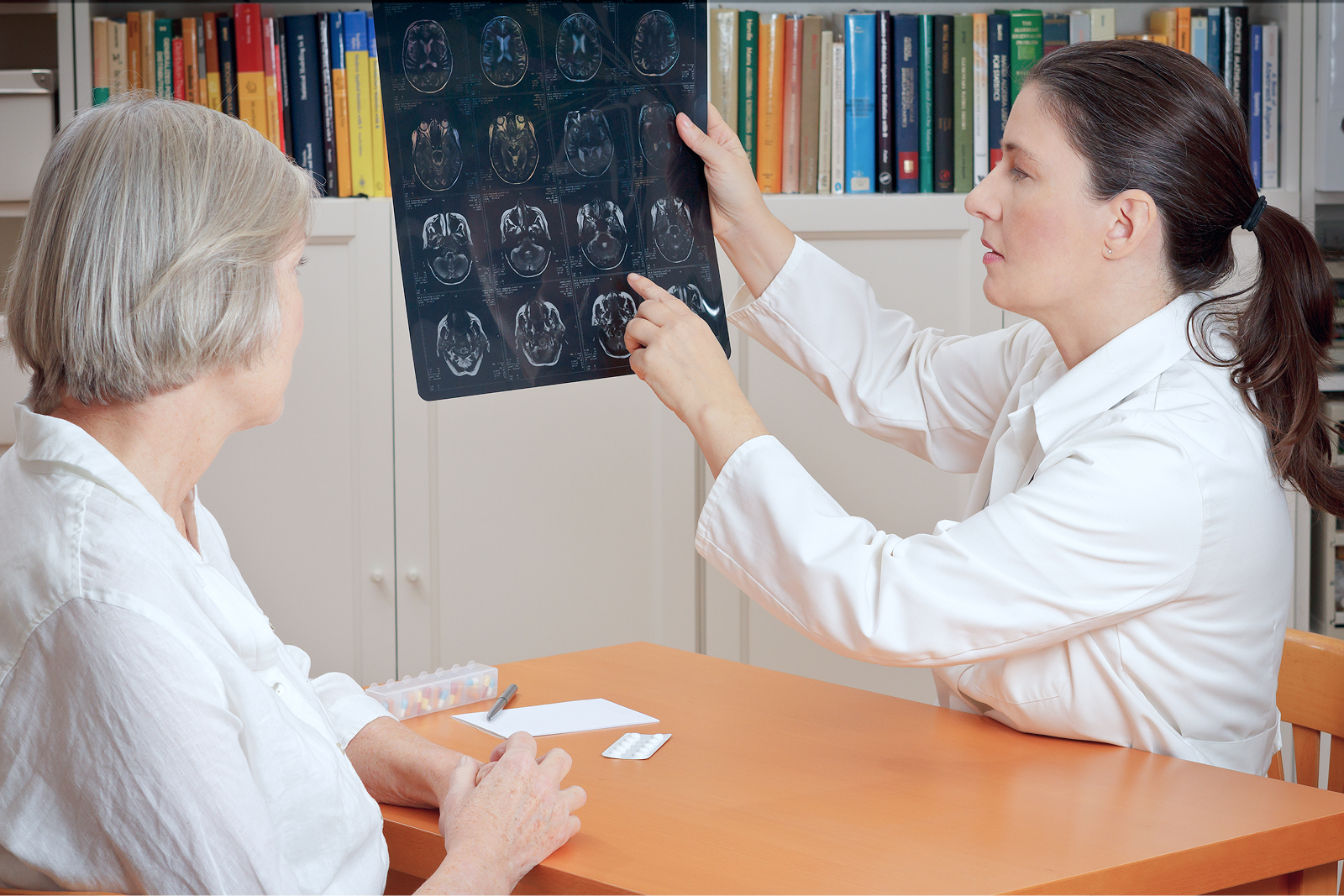 Ärztin zeigt einer Patientin Röntgenbilder, während sie an einem Tisch in einem Raum mit Bücherregalen sitzen.