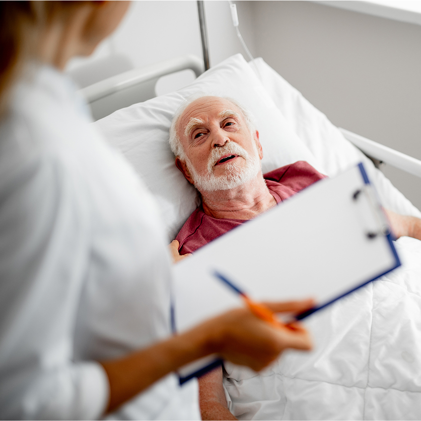 Ein älterer Mann liegt im Krankenhausbett und spricht mit einer Ärztin, die ein Klemmbrett hält und sich Notizen macht.