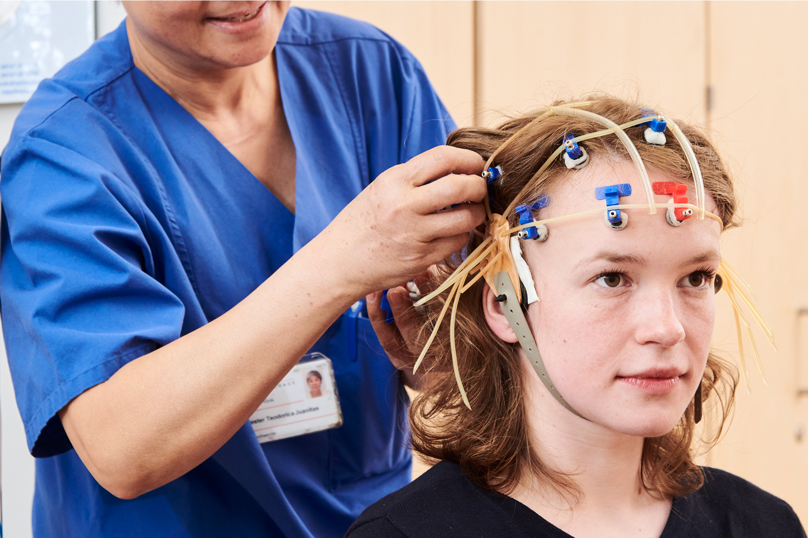 Eine junge Person sitzt, während ihr eine andere Person in blauer Arbeitskleidung Elektroden am Kopf anbringt.