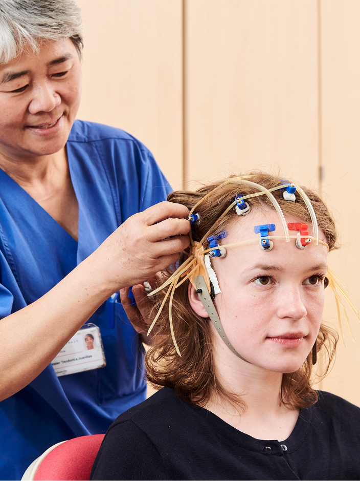 Eine junge Person mit einem EEG-Haarband wird von einer Fachkraft in blauer Kleidung für eine Untersuchung vorbereitet.