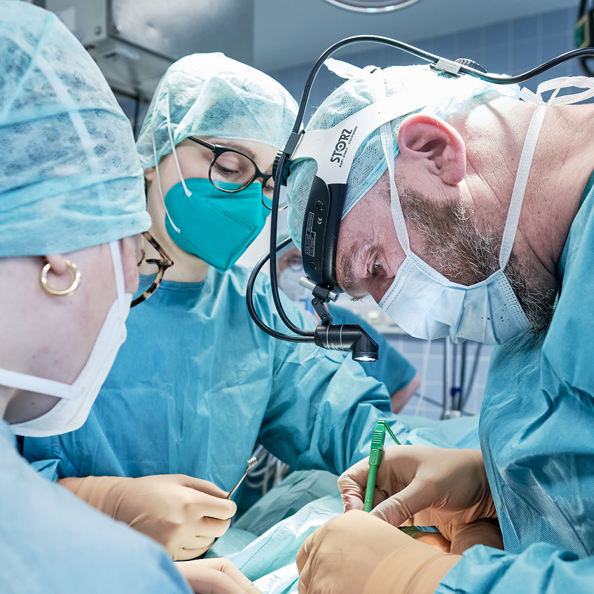 Drei Chirurgen in OP-Kleidung führen eine Operation durch, konzentriert über den Patienten vor ihnen gebeugt.