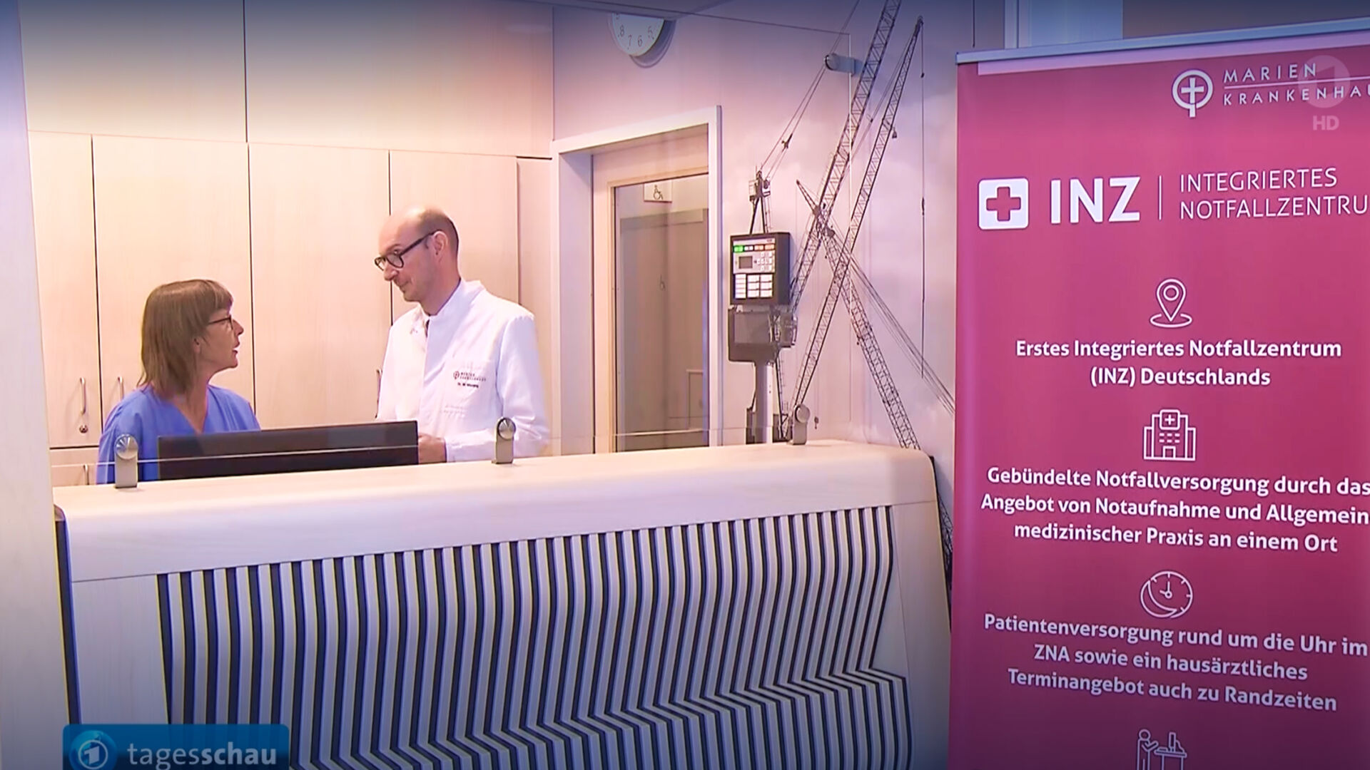Zwei Personen stehen hinter einem Empfangstresen, im Vordergrund steht ein Informationsbanner mit Hinweisen zum INZ.