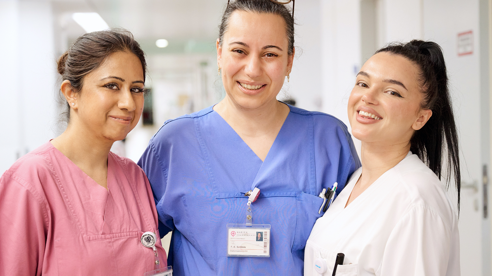 Drei Frauen in medizinischer Kleidung stehen zusammen in einem Krankenhausflur und lächeln in die Kamera.