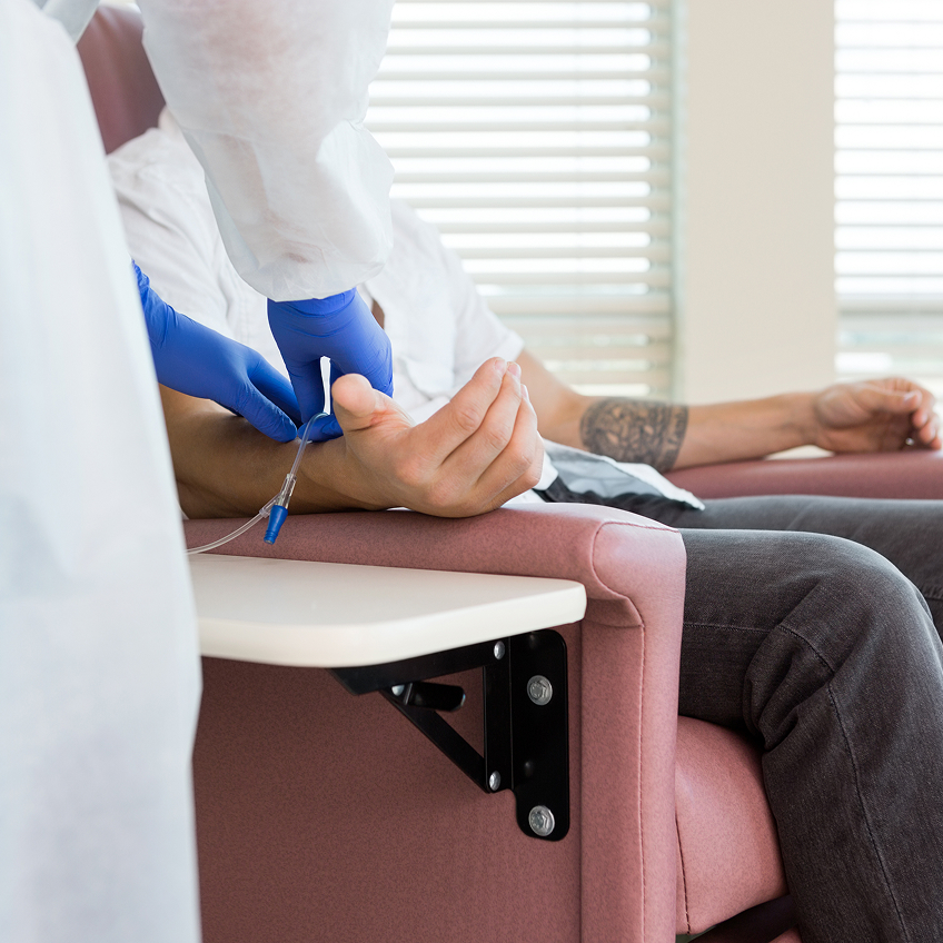Eine Person sitzt in einem Stuhl, während eine andere Person ihr einen Zugang am Arm legt.
