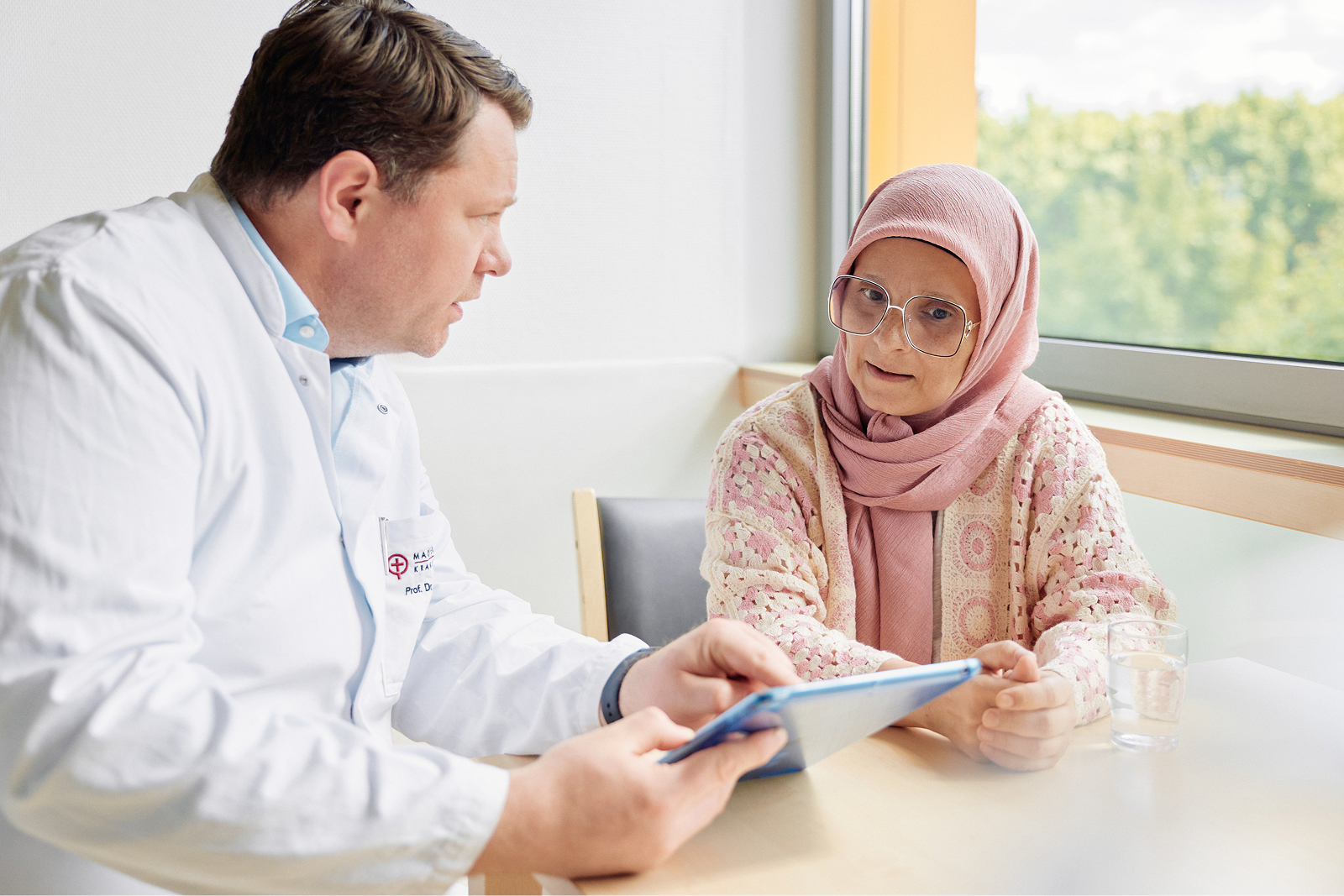 Ein Arzt und eine Patientin im Gespräch, während sie ein Tablet betrachten. Die Patientin trägt ein Kopftuch.