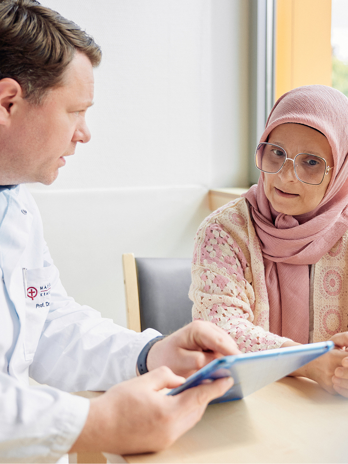 Ein Arzt und eine Patientin im Gespräch, während sie ein Tablet betrachten. Die Patientin trägt ein Kopftuch.