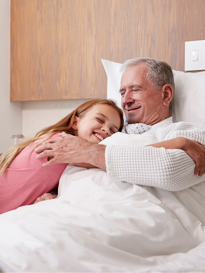 Ein älterer Mann sitzt im Krankenhausbett, während er ein lächelndes Mädchen zur Begrüßung umarmt.