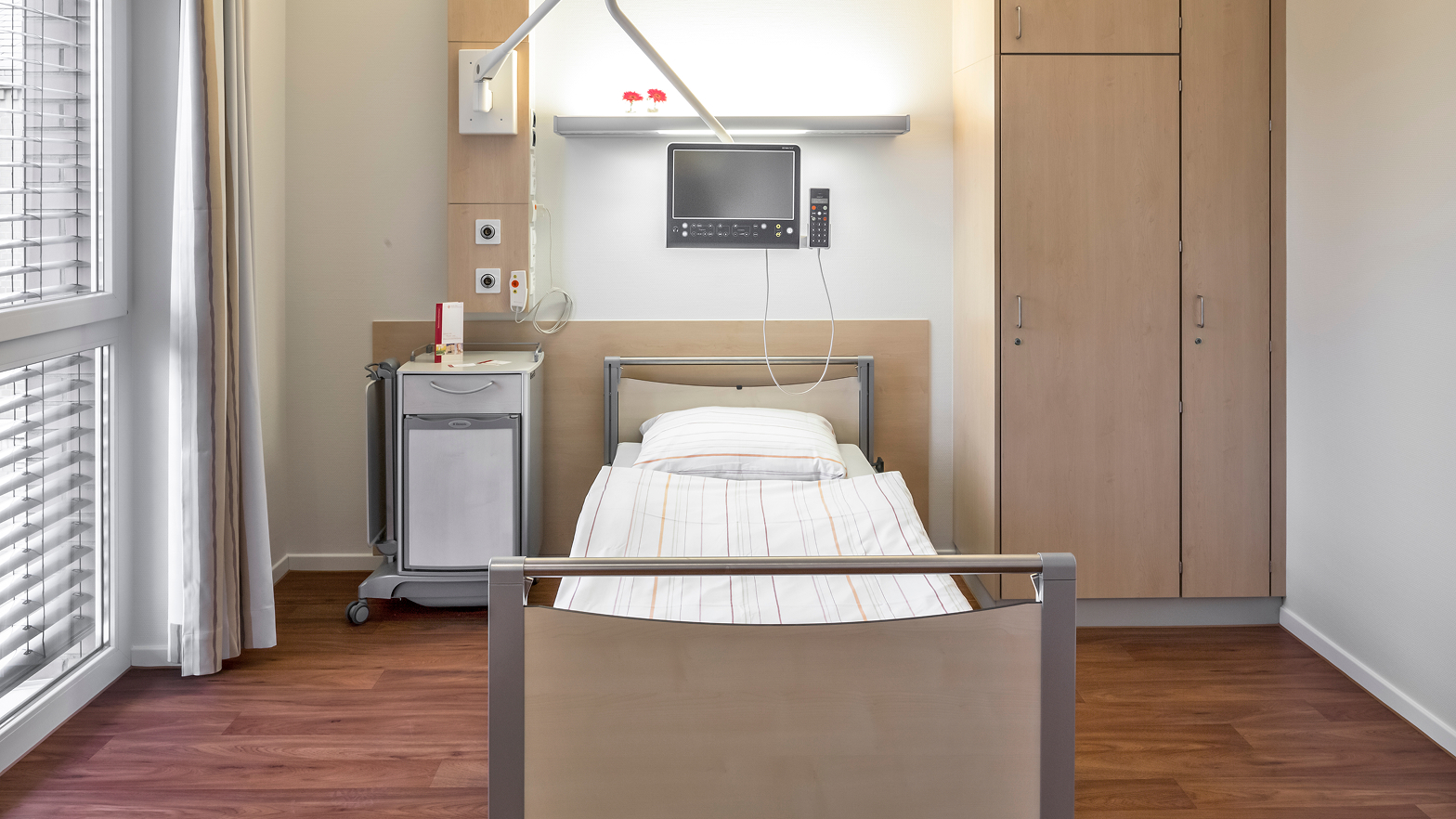 Ein modernes, helles Krankenhauszimmer mit einem Bett, einem Schrank, einem Nachttisch und einem Monitor an der Wand.