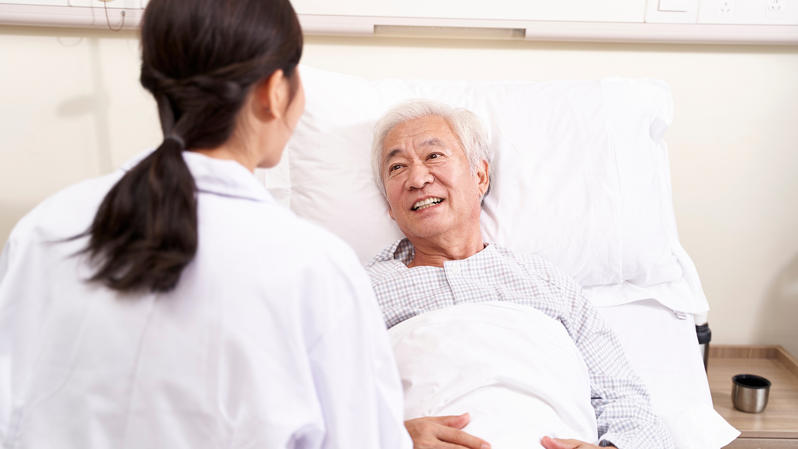 Ein älterer Mann liegt im Krankenhausbett und lächelt eine dunkelhaarige Ärztin an, die vor ihm steht.