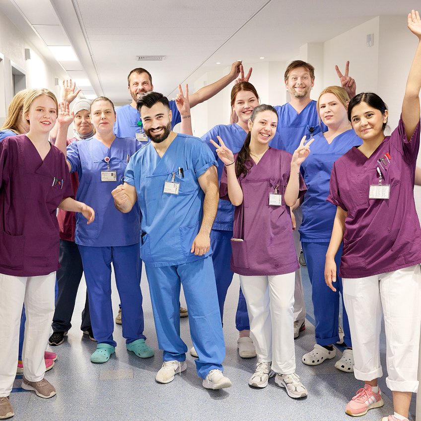 Gruppe von medizinischem Personal in blauer und lilafarbener Arbeitskleidung posiert fröhlich in einem Klinikflur.