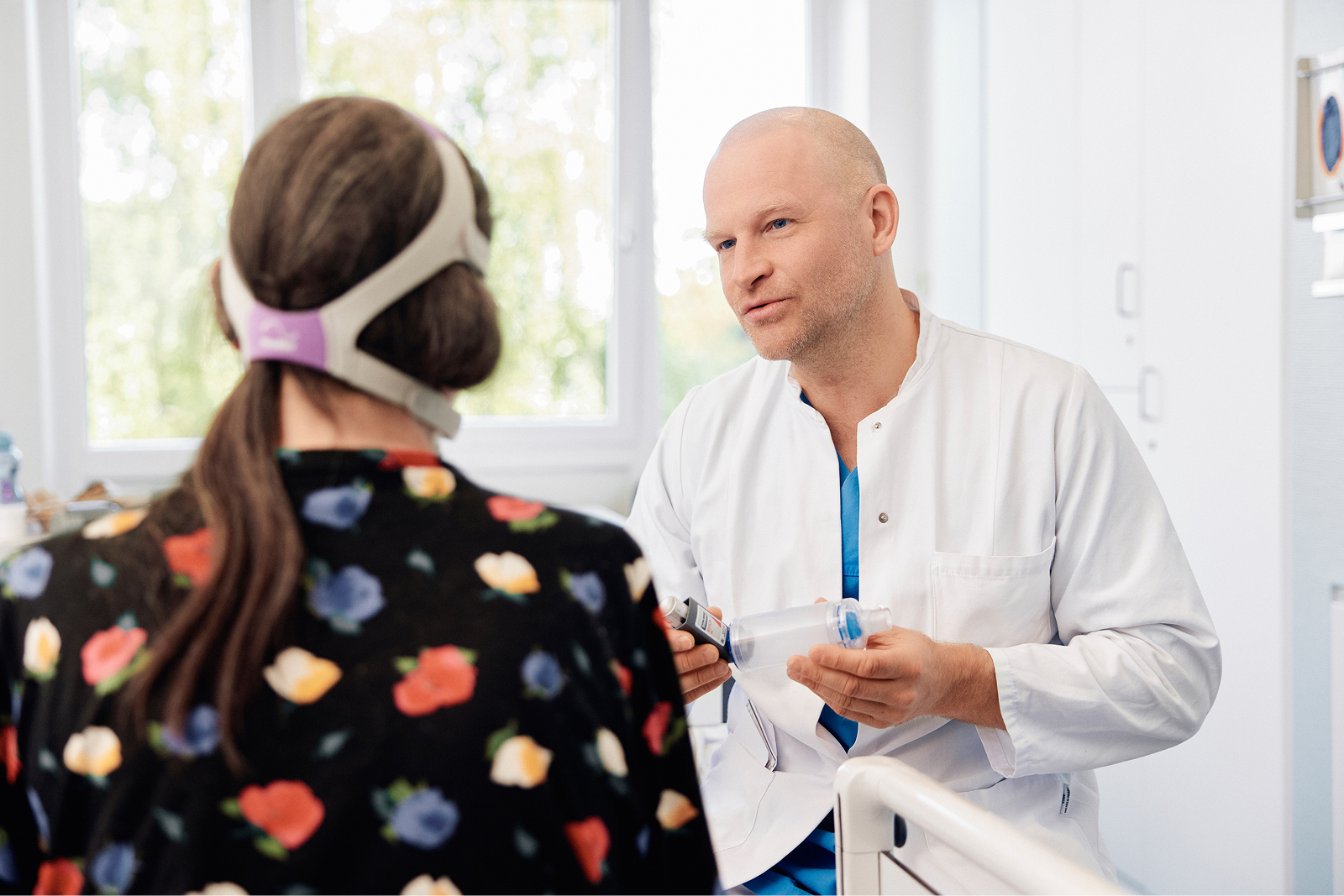 Ein Arzt spricht in einem hellen Krankenzimmer mit einer Patientin, die eine Atemmaske trägt.