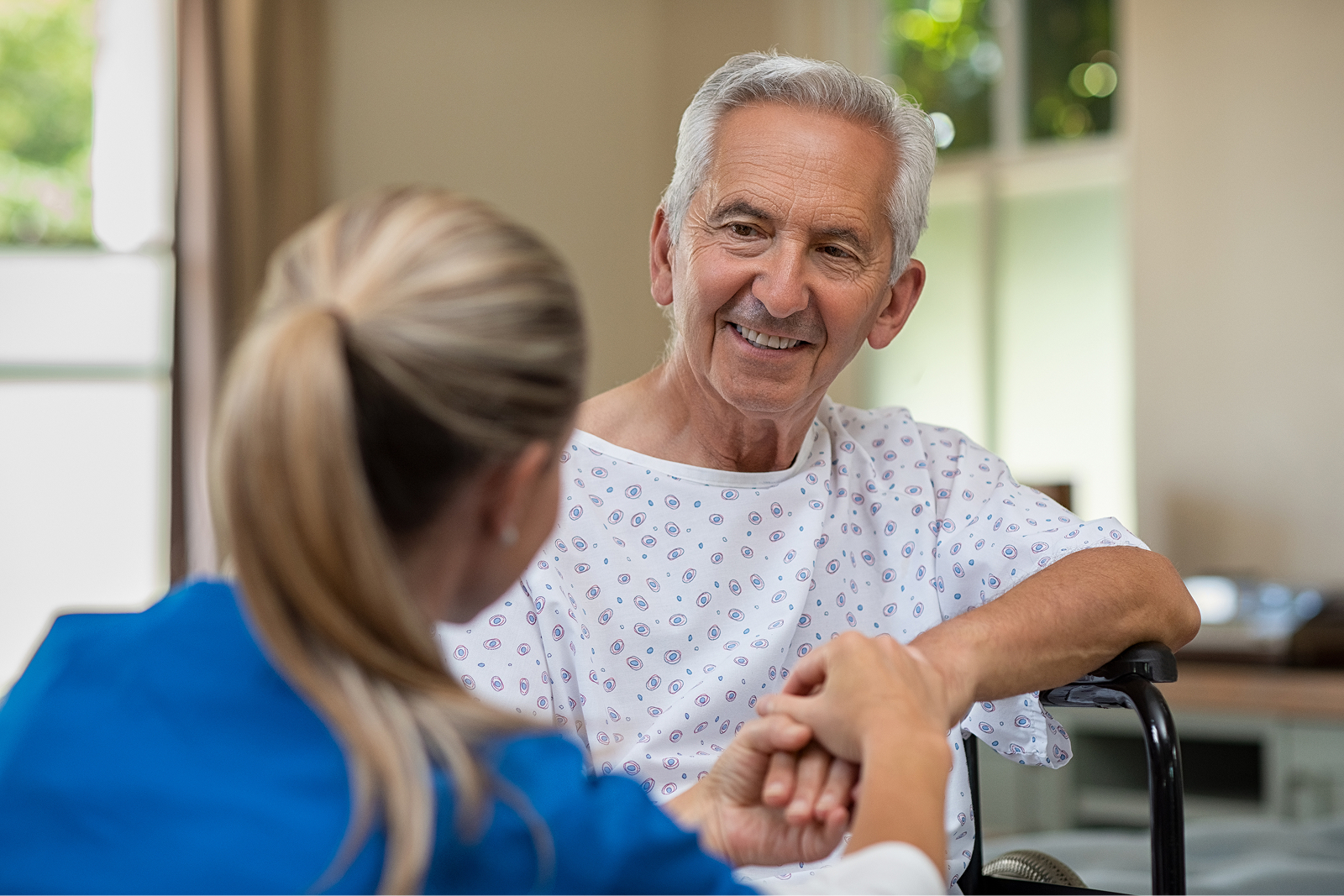 Ein älterer Mann in einem Krankenhaushemd lächelt einer Pflegekraft zu, die ihm die Hand hält.