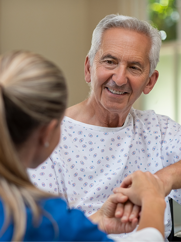 Ein älterer Mann in einem Krankenhaushemd lächelt einer Pflegekraft zu, die ihm die Hand hält.