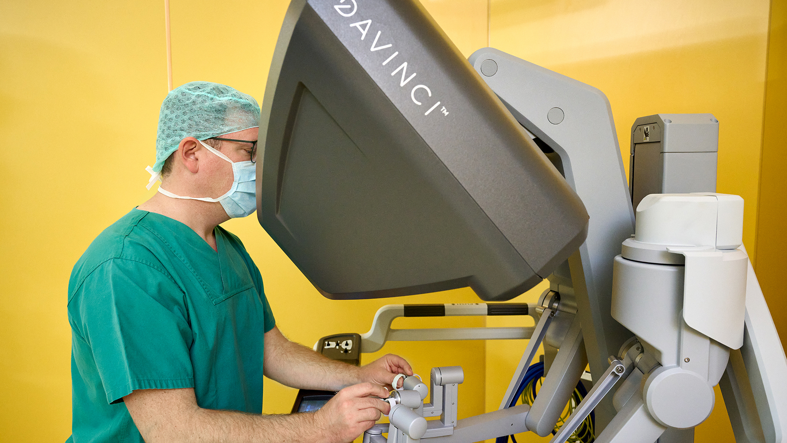 Ein Chirurg in grüner Kleidung bedient ein Da Vinci-Operationssystem vor einer gelben Wand.