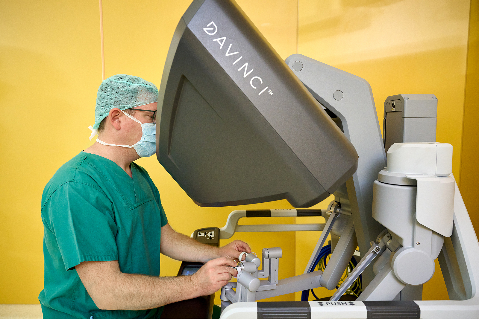 Ein Arzt in grüner OP-Kleidung bedient ein Da Vinci-Operationssystem in einem gelben Raum.