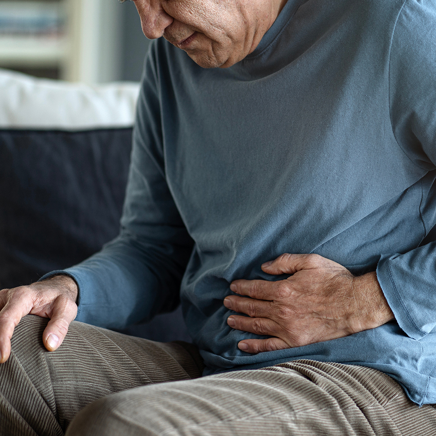 Älterer Mann im Blauen Pullover sitzt auf einem Sofa und hält sich mit einer Hand den Bauch.