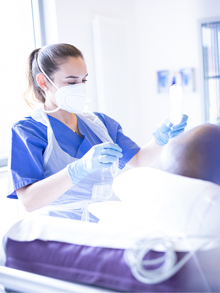 Eine medizinische Fachkraft mit blauer Arbeitskleidung und Mundschutz kümmert sich um einen Patienten.